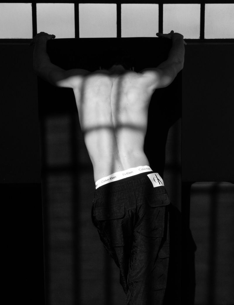 Monochromatic shot of muscular shirtless man in dark trousers leaning against paneled structure with light and shadow play on back.