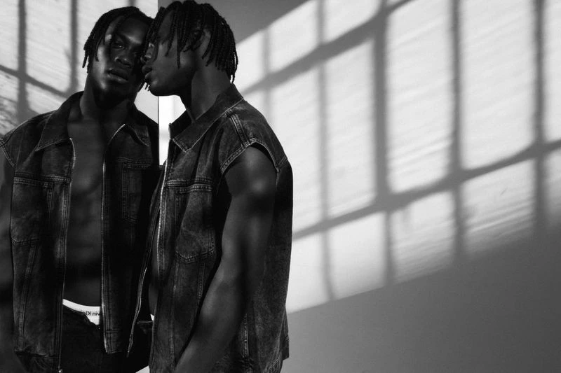 Striking black and white portrait of an African American male model in a denim vest, leaning against a reflective surface with shadow patterns.