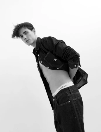 A young adult man poses indoors, wearing dark denim shirt and jeans, with Calvin Klein branded underwear, in a black and white studio shot.