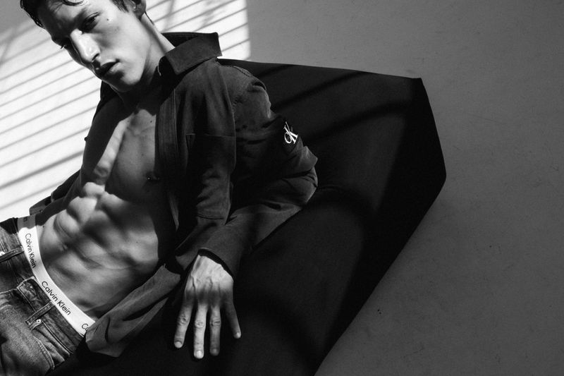 Black and white studio shot of a muscular adult male lying down wearing jeans and a jacket revealing his abs, with window blinds.
