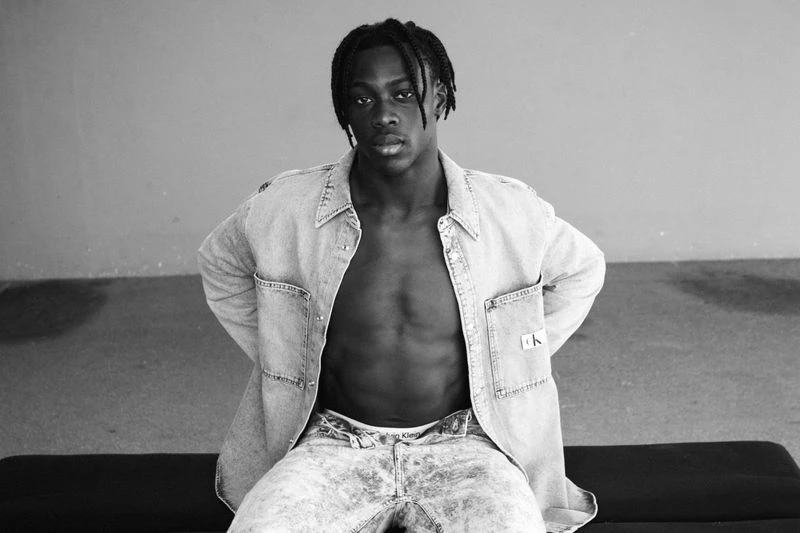 Black and white portrait of a young Black adult male with an open denim shirt, acid-wash jeans, sitting indoors against a grey wall.