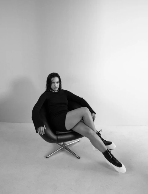 Woman in black dress and chunky boots sits in a modern chair against a grey background, fashion studio shot.