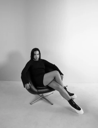 Woman in black dress and chunky boots sits in a modern chair against a grey background, fashion studio shot.
