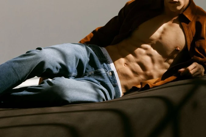 Athletic man in jeans and an open brown shirt, reclining on a brown surface, showcasing abdominal muscles, wearing Calvin Klein underwear.