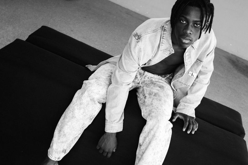 Black and white portrait of a young man in a denim jacket and jeans sitting on a black surface, looking toward the camera.