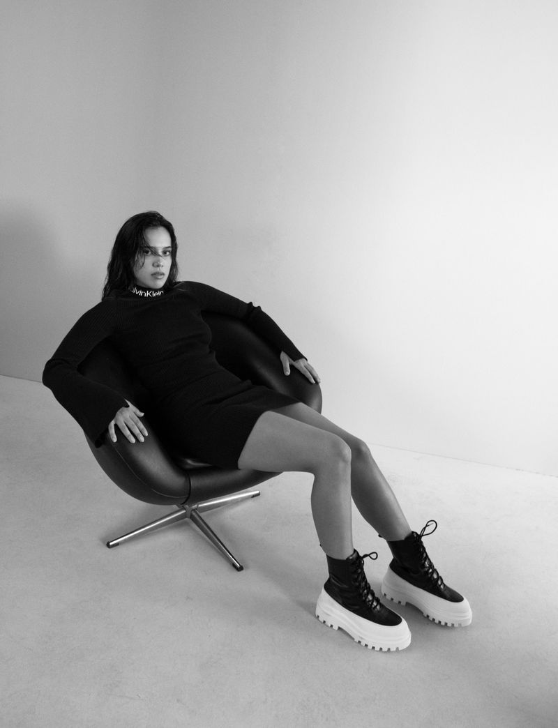 Monochrome portrait of a woman sitting in a black chair, wearing a black dress and chunky heeled black and white boots in studio lighting.
