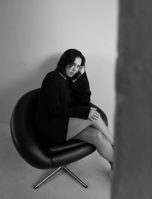 A woman in a black dress is seated in a black chair in a studio. Black and white portrait featuring fashion, and indoor setting.