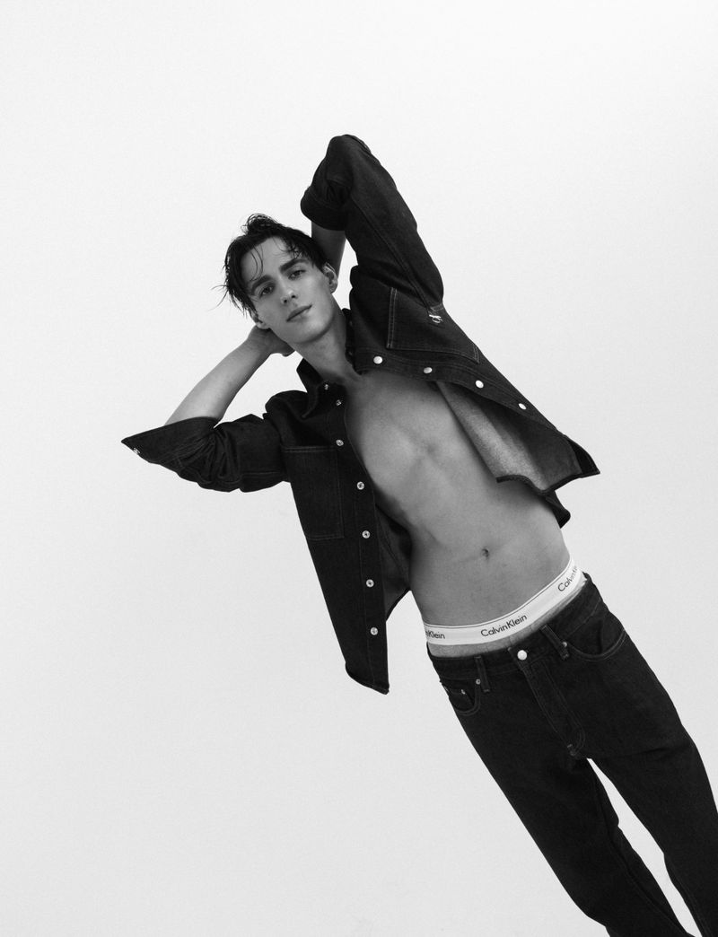 Black and white portrait of a male model wearing an unbuttoned denim shirt and jeans against a white background