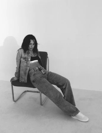 Monochrome fashion image of a model wearing denim jacket and jeans seated on a tubular chair against a white wall