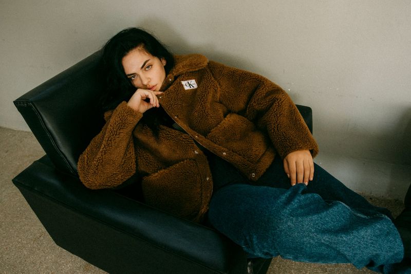 A young woman with dark hair poses in a studio armchair, wearing a brown sherpa jacket and jeans, against a neutral gray background.