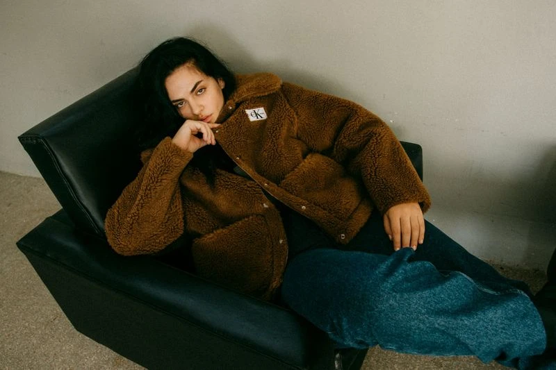 A young woman with dark hair poses in a studio armchair, wearing a brown sherpa jacket and jeans, against a neutral gray background.