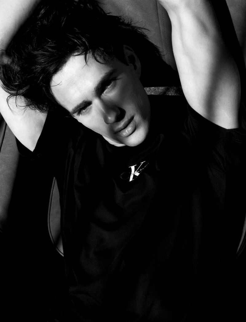 Black and white portrait of model Iasonas Laios in a black Calvin Klein t-shirt reclining with arms raised overhead.