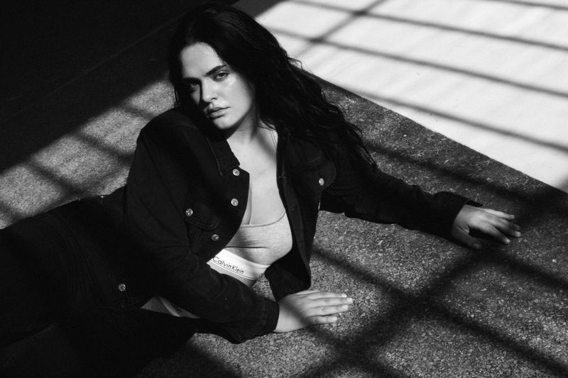 Portrait of a woman with dark hair wearing a denim jacket and underwear, lying on the floor with strong window-shaped shadows.
