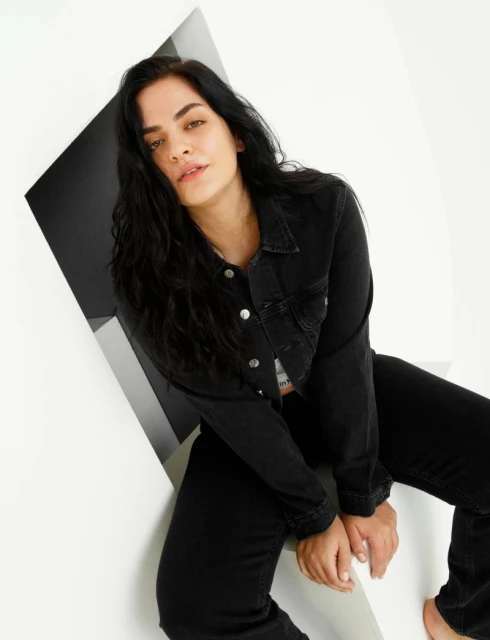 A woman with dark hair poses in black denim jacket and pants against a geometric white and gray backdrop in a studio setting.
