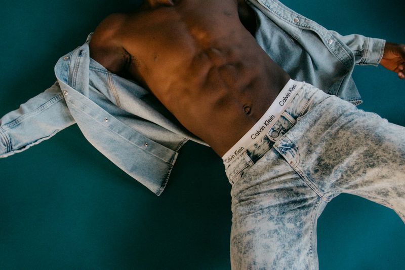 A Black male model is lying against a teal background while wearing jeans and a denim shirt that is falling off his shoulders.