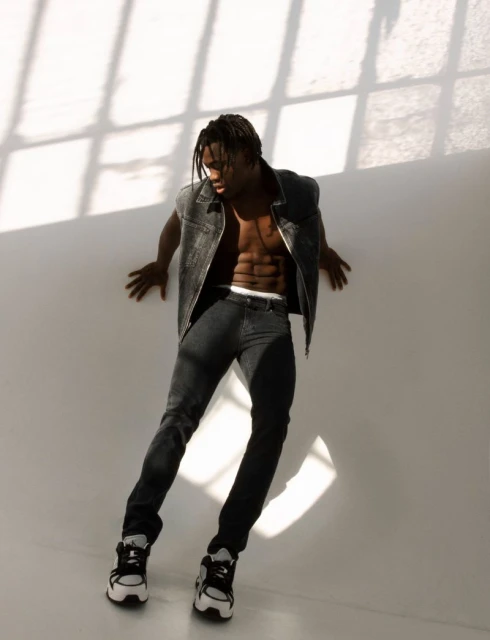 Athletic Black man with bare torso poses indoors against a white wall wearing a vest, black pants and sneakers, with window shadow.