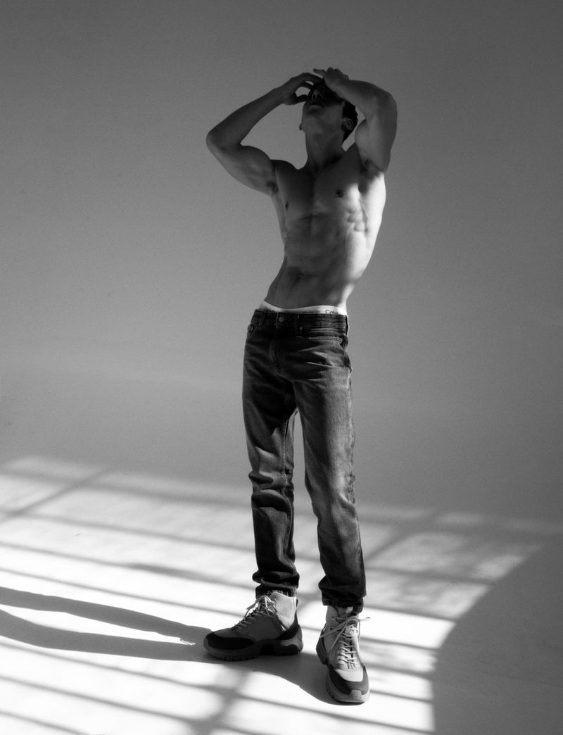Shirtless male model wearing dark denim jeans and boots posing with hands on head in a studio