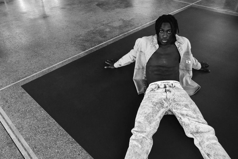 A young Black male is laying on a black and grey marble floor wearing a denim jacket and jeans, posing with an open torso.
