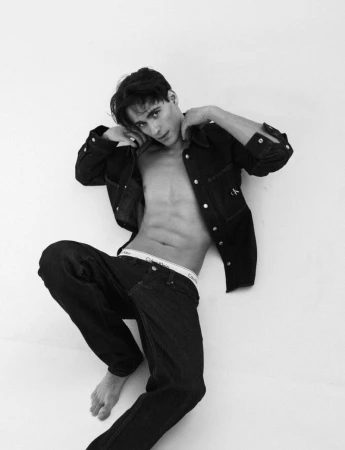 Iasonas Laios lying barefoot in an unbuttoned black denim jacket, matching jeans, and white waistband on a white background.