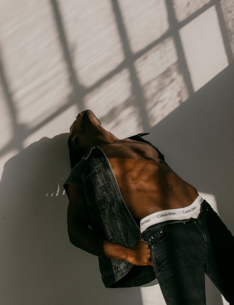 An adult male in black denim and white underwear reclines against a wall with window shadows. Indoor fashion shot, close up.