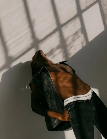 An adult male in black denim and white underwear reclines against a wall with window shadows. Indoor fashion shot, close up.