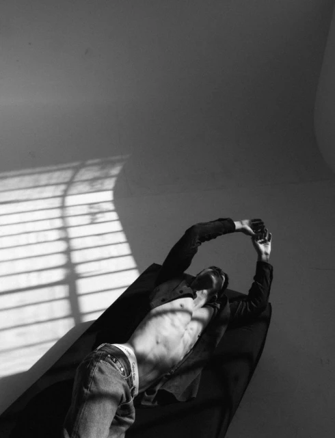 Black and white studio shot of a muscular man lying on a dark surface in blue jeans and an unbuttoned jacket, posing with arms overhead.