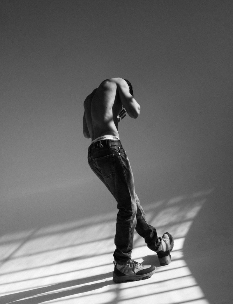 Black and white portrait of a shirtless man wearing jeans, posing in a photo studio with sunlight streaming through the blinds.