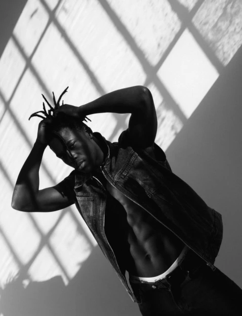 Striking black and white portrait of a Black male model with dreadlocks posing in denim vest with window shadow patterns on the backdrop.