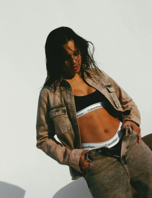 A woman poses in a light brown denim jacket and jeans, wearing Calvin Klein branded underwear against a neutral backdrop.