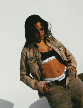 A woman poses in a light brown denim jacket and jeans, wearing Calvin Klein branded underwear against a neutral backdrop.