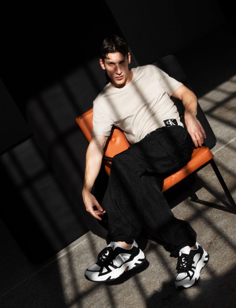 Man in tee shirt and black pants sits on an orange chair. He wears white sneakers in a photo studio with shadows.
