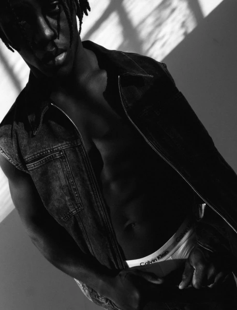 Black and white shot of a Black male model posing with an open denim vest, Calvin Klein underwear, and studio lighting with shadow patterns.