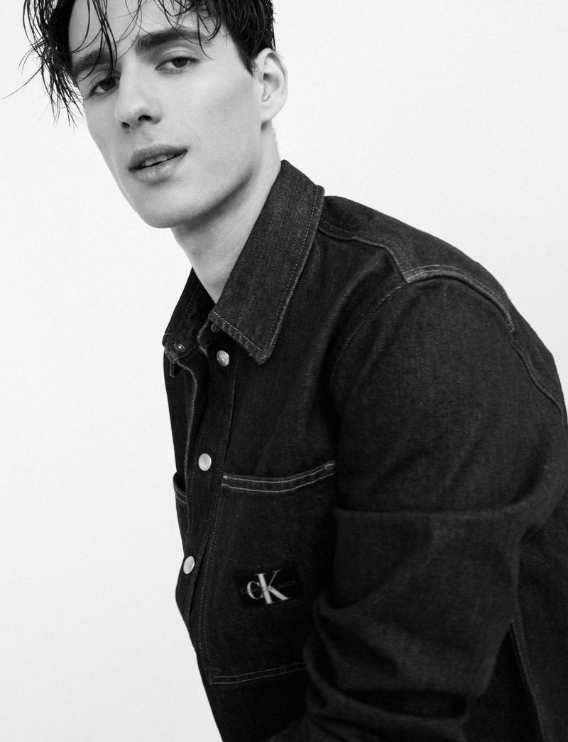 Monochromatic portrait of a young adult male wearing a dark denim jacket with a visible logo, posing against a bright white backdrop.