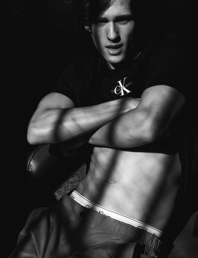 Black and white portrait of a young man with his arms crossed, wearing a Calvin Klein t-shirt and shorts in a shadowed setting.