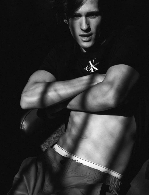 Black and white portrait of a young man with his arms crossed, wearing a Calvin Klein t-shirt and shorts in a shadowed setting.