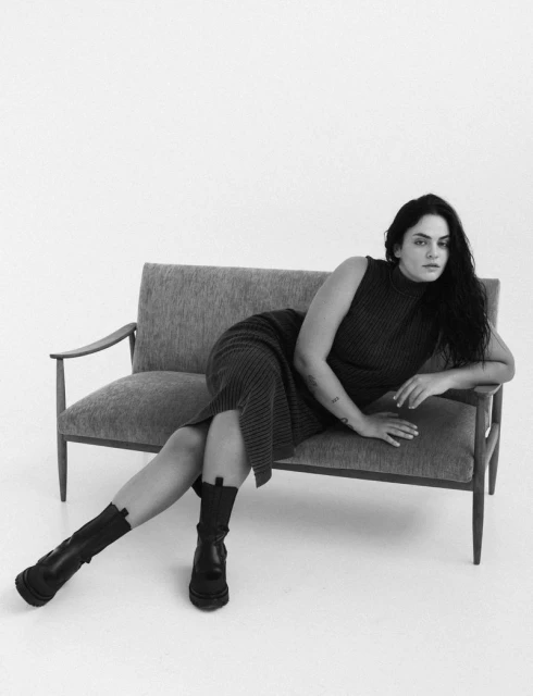 A woman with long dark hair poses on a gray couch in a sleeveless sweater dress and black boots against a white backdrop in a studio.