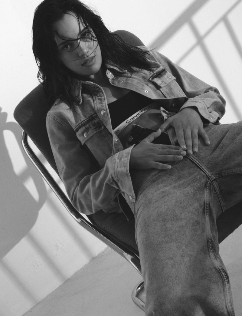Black and white portrait of a woman in denim jacket and jeans sitting in a chair with light and shadow falling on the wall.