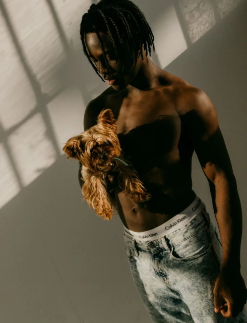 Man holding a dog in an indoor studio, bathed in window light with shadow patterns and wearing jeans with a Calvin Klein branded waistband.