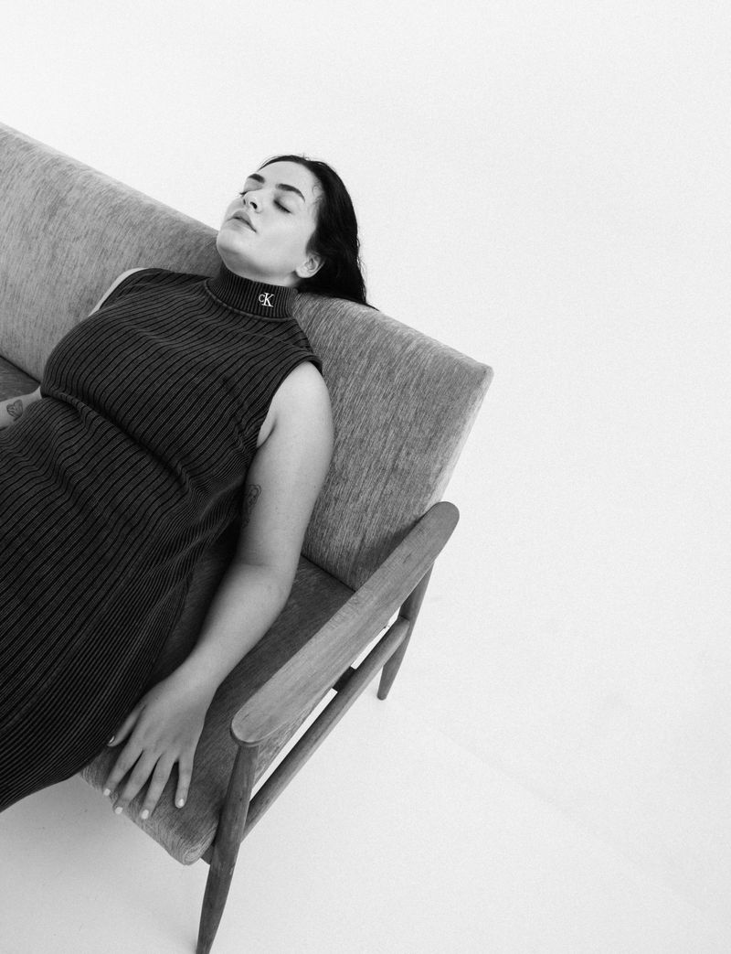 Woman rests on a modern grey sofa in black and white. She wears a ribbed, sleeveless dress and relaxes against a white backdrop.