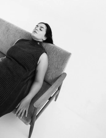 Woman rests on a modern grey sofa in black and white. She wears a ribbed, sleeveless dress and relaxes against a white backdrop.