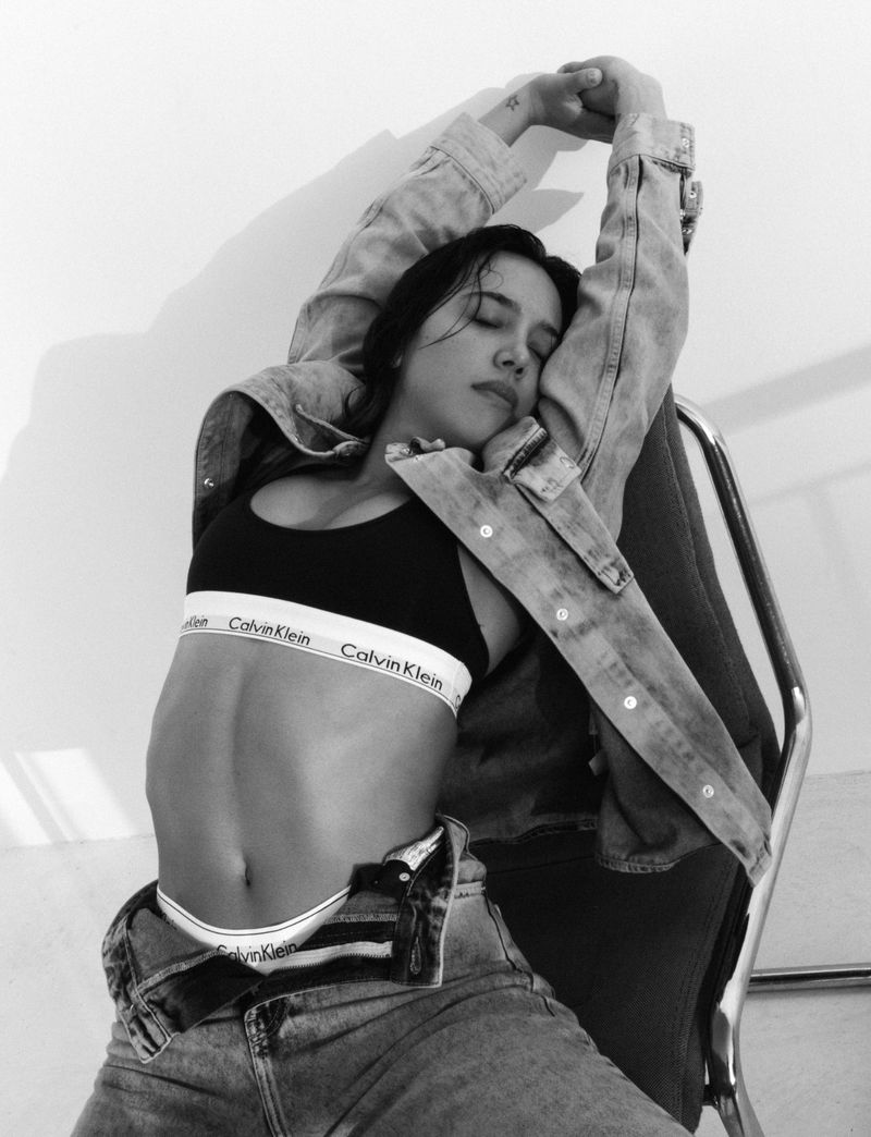 Monochrome portrait of a woman with closed eyes, wearing a bra top and jeans, leaning back in a chair in a photo studio.