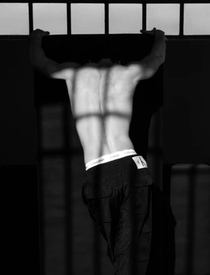 Black and white photo of a shirtless man viewed from behind in black trousers and Calvin Klein underwear with window shadows across his back.