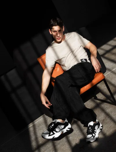 Model Iasonas Laios seated in an orange armchair wearing a beige t-shirt and black pants under sharp grid shadows.
