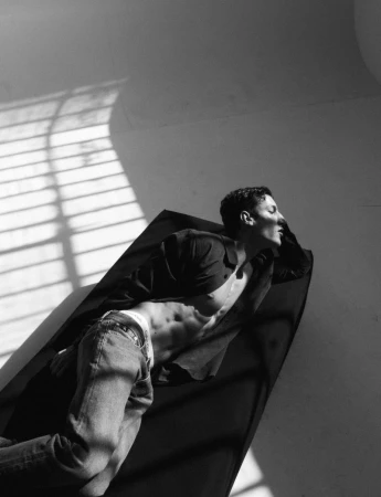 Model Iasonas Laios reclining shirtless in an open jacket and jeans under dramatic window shadows.