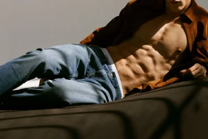 Model Iasonas Laios reclining in blue denim jeans, white underwear waistband, and an open brown overshirt.