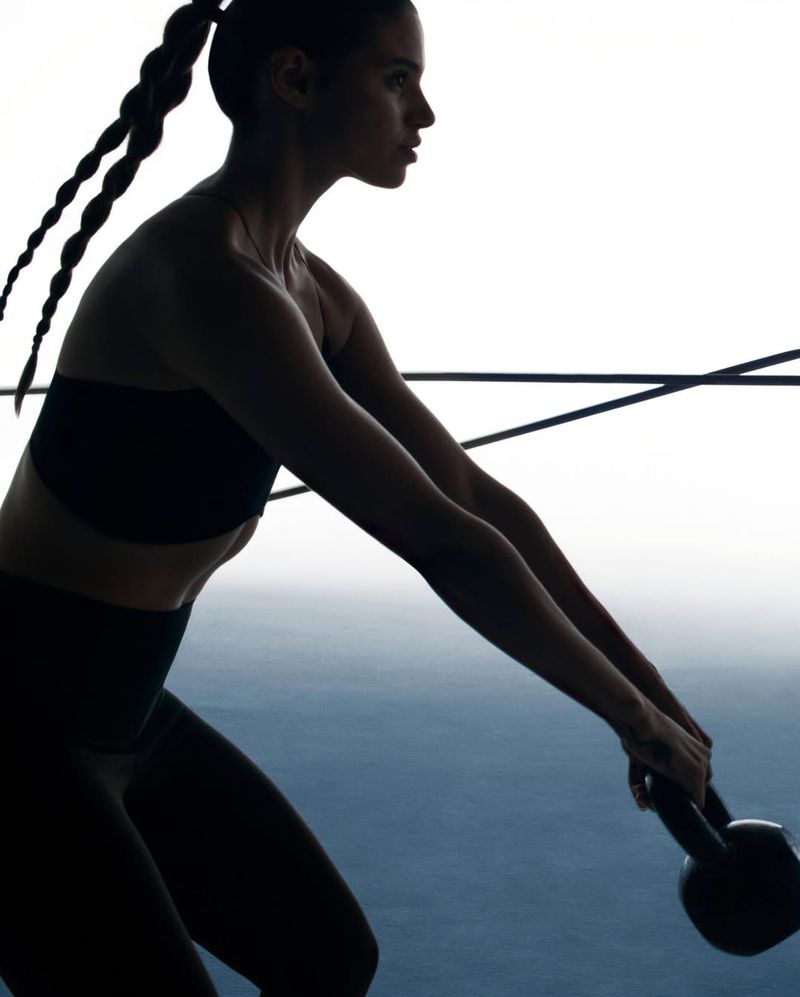 Silhouette of woman in fitness clothing lifting kettlebell in gym setting; black and white with backlighting and minimalist composition.