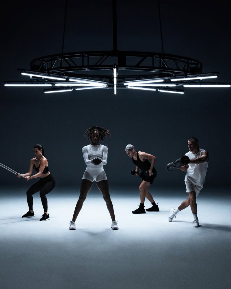 Four people are exercising in a studio. One person is using a jump rope, one is doing squats, another is lifting weights, and one is holding a sandbag.