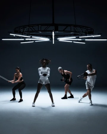 Four people are exercising in a studio. One person is using a jump rope, one is doing squats, another is lifting weights, and one is holding a sandbag.