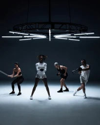Four athletes in monochrome activewear training beneath an overhead circular lighting rig in a dark studio.