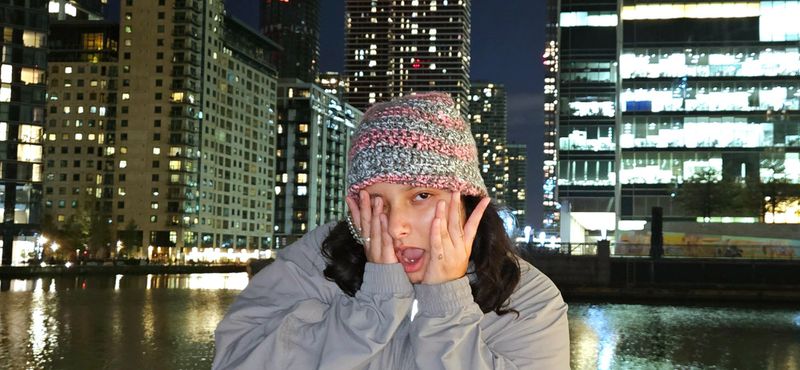 Night shot of a person wearing a hat, standing in front of a body of water, with a backdrop of illuminated city skyscrapers.